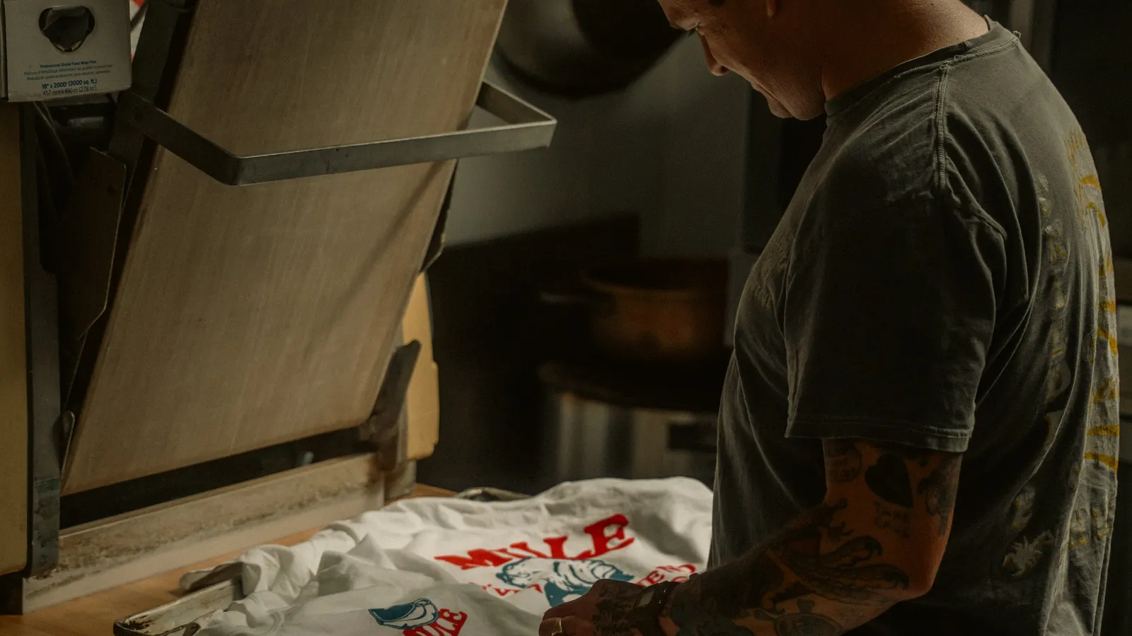Operador preparando camiseta ao lado de uma prensa térmica em oficina de personalização